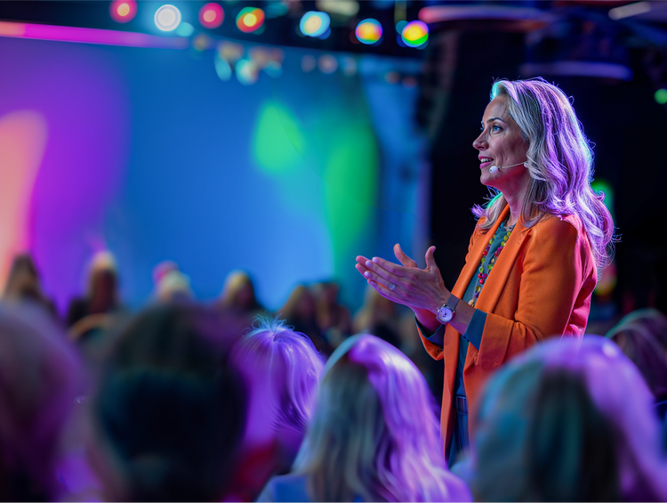 Female speaker in orange blazer presenting at marketing conference with purple stage lighting